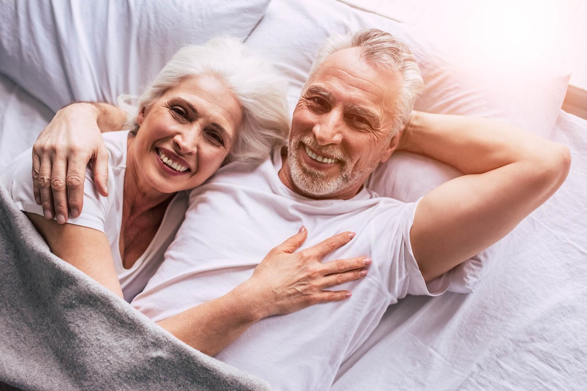 A senior couple cuddled up in bed smiling.